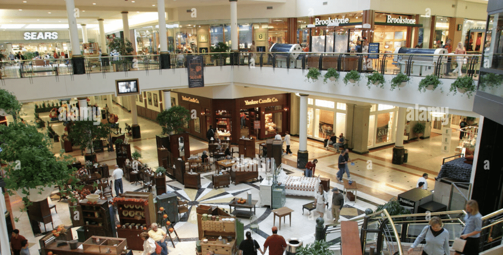 A multi-level shopping mall interior with people walking, various stores including Sears and Brookstone, indoor plants, and display kiosks in the central area.